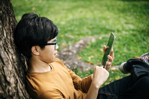 молодий чоловік використовує планшет і сидить під деревом
 - Фото, зображення