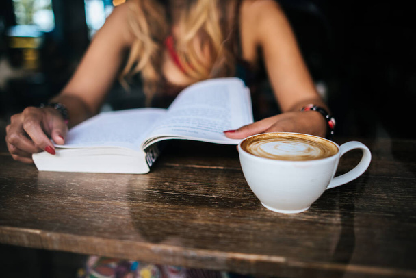 Νεαρή γυναίκα διαβάζει βιβλίο και φρέσκο φλιτζάνι καφέ στο τραπέζι - Φωτογραφία, εικόνα
