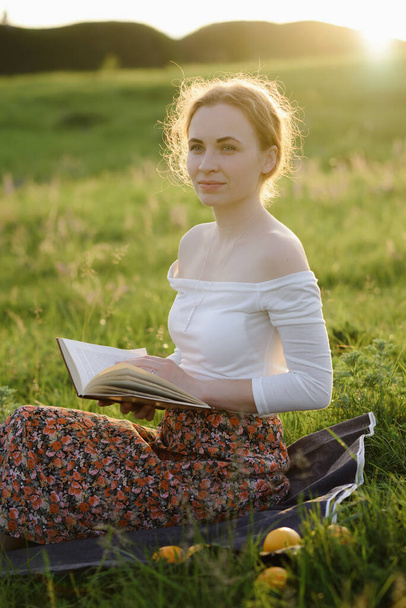 Νεαρό κορίτσι διαβάζει ένα βιβλίο στο γρασίδι, ενώ δύει ο ήλιος. Καλοκαιρινή αίσθηση - Φωτογραφία, εικόνα