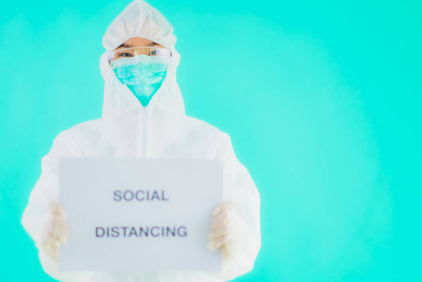 Portrait beautiful young asian doctor woman wear ppe or  personal protective equipment for protect from coronavirus or covid19 with social distacing sign on blue isolated background - Photo, Image