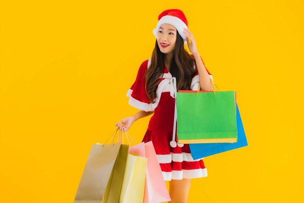 Porträt schöne junge asiatische Frau tragen Weihnachtskleidung Hut mit einer Menge Einkaufstasche auf gelbem Hintergrund isoliert - Foto, Bild