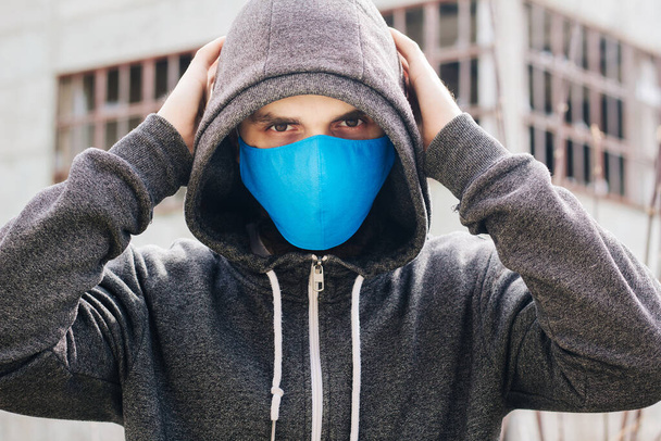 Chlap v šedém svetru s modrou lékařskou maskou. Drží kapuci vlastníma rukama. Na pozadí zničených a opuštěných budov. Muž v karanténě. - Fotografie, Obrázek