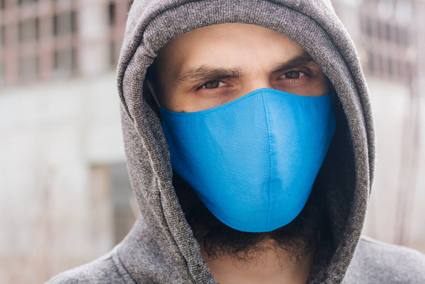 A guy in a gray sweater with a blue medical mask. Holds a hood with his own hands. Against the background of destroyed and abandoned buildings. Quarantined man. - Valokuva, kuva