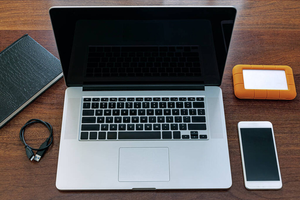 Άποψη της μαύρης οθόνης του laptop, μια συσκευή για να εργαστείτε στο σπίτι, να κάνετε έρευνα, ατζέντα, HD, κινητό τηλέφωνο και USB drive. Πάνω από ένα ξύλινο τραπέζι γραφείου. - Φωτογραφία, εικόνα