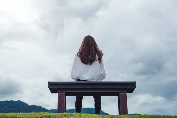 Een vrouw zit alleen op een houten bank in het park met bewolkte en sombere lucht achtergrond - Foto, afbeelding