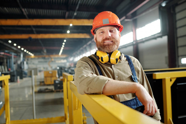 Портрет позитивного працівника чоловічої статі в помаранчевому хардхаті, що спирається на металеві перила в сучасному заводі
 - Фото, зображення