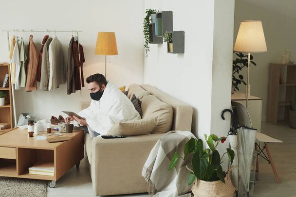 Kranker junger Mann mit schwarzer Maske sitzt auf der Couch und benutzt Touchpad, während er in der Isolation zu Hause fernarbeitet - Foto, Bild