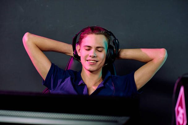 Молодий усміхнений розслаблений відеогравець чоловічої статі в навушниках насолоджується перервою між електронними спортивними іграми, тримаючи руки за голову
 - Фото, зображення