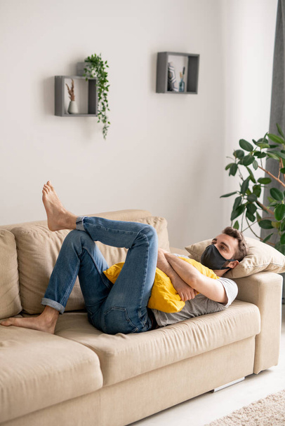 Σοβαρός νεαρός άνδρας με προστατευτική μάσκα ξαπλωμένος με μαξιλάρι στον καναπέ και αίσθημα κατάθλιψης λόγω κοινωνικής απομόνωσης - Φωτογραφία, εικόνα