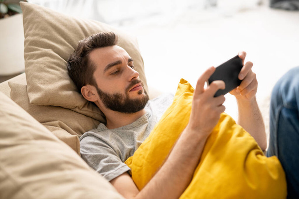 Jeune homme barbu sérieux avec un oreiller jaune couché sur le canapé et regardant une vidéo internet sur smartphone en quarantaine - Photo, image