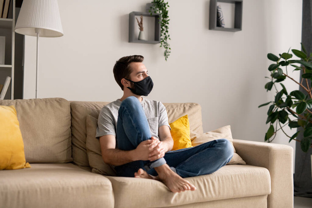 Συλληπτικός νεαρός άνδρας με υφασμάτινη μάσκα, απομονωμένος στο σπίτι, κάθεται στον καναπέ και κοιτάζει αλλού. - Φωτογραφία, εικόνα