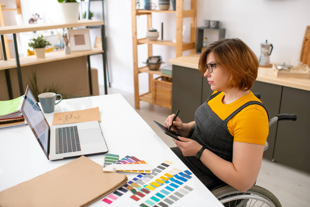 Evde tasarım projesi üzerinde çalışırken tekerlekli sandalyede oturan ve grafik tasarım tableti çizen meşgul genç bir kadın. - Fotoğraf, Görsel