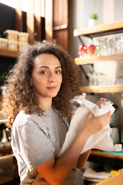 Pretty young waitress or restaurant worker in t-shirt and apron looking at you while washing glasses and wiping them by workplace - Photo, Image