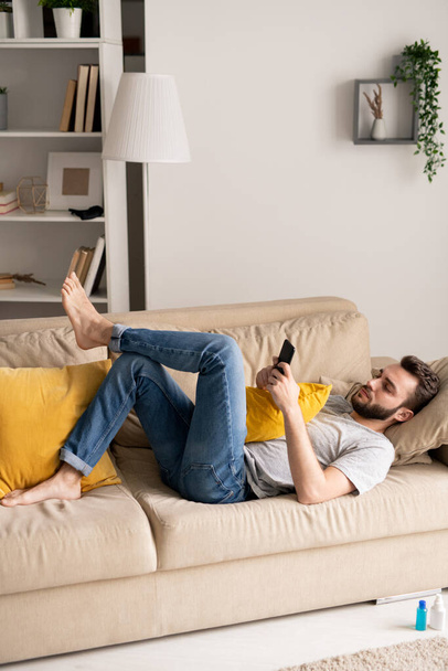Vážný mladý vousatý muž ležící na pohodlné pohovce a pomocí smartphone v domácí izolaci - Fotografie, Obrázek