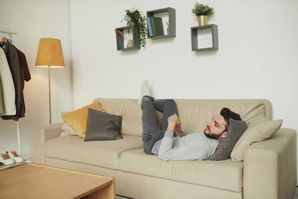 Ξεκούραστος άντρας με καθημερινά ρούχα ξαπλωμένος στον καναπέ με το κεφάλι του σε μαξιλάρια μπροστά στην τηλεόραση και βλέποντας ταινία ή μεταδίδοντας στο σαλόνι - Φωτογραφία, εικόνα