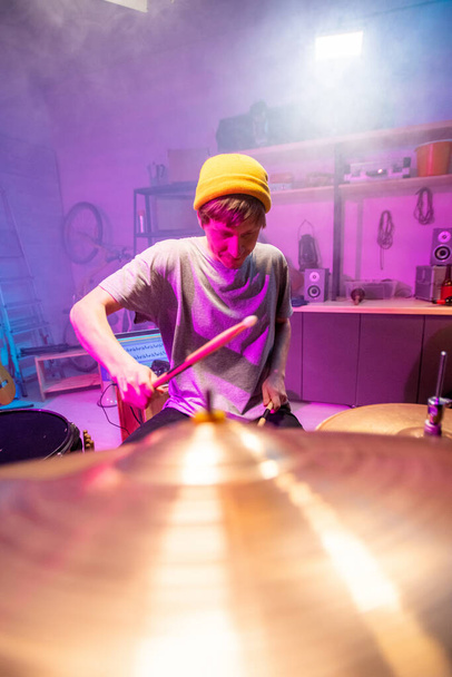 Joven músico guapo inclinándose sobre batería mientras golpea tambores y platillos y la grabación de música para una nueva canción en el ambiente del garaje
 - Foto, Imagen