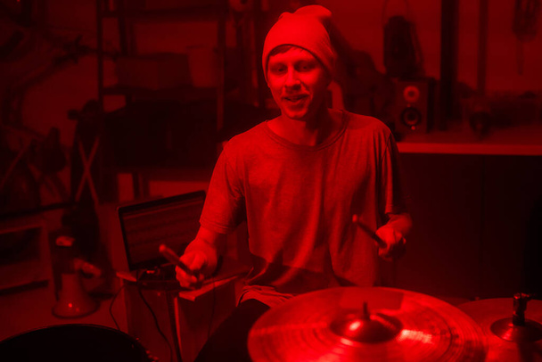 Молодой музыкант в шапочке и футболке стучит барабанными палочками по тарелкам и барабанам, сидя за барабанной установкой и ноутбуком в гараже
 - Фото, изображение