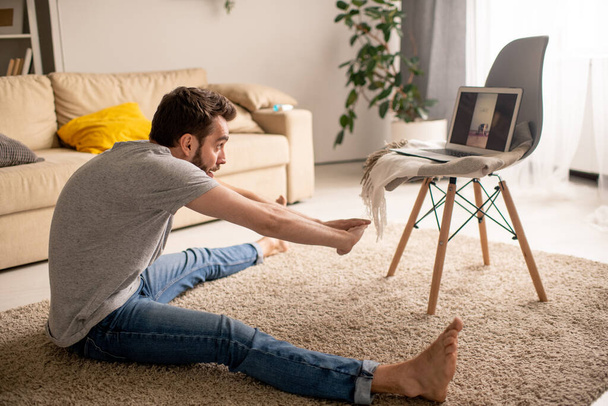 Jeune homme inflexible en vêtements décontractés assis sur le tapis et pratiquant grand angle assis en avant courbure tout en regardant cours de yoga en ligne - Photo, image