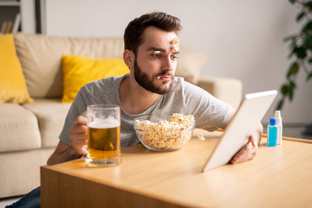Śpiący młody brodaty mężczyzna z popcornami na twarzy pijący piwo i oglądający film na tablecie, leniwy dzień w kwarantannie - Zdjęcie, obraz