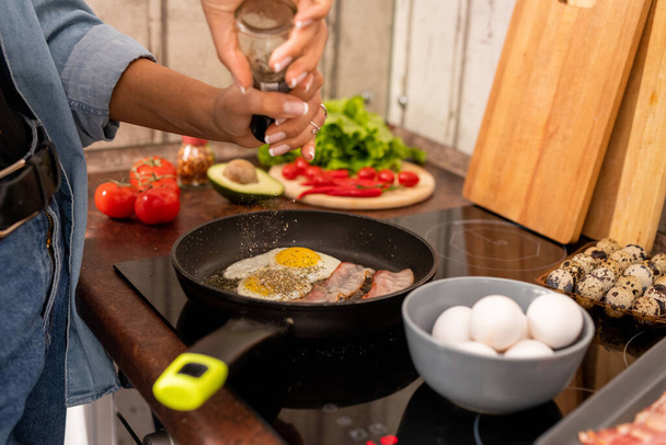 Hands of young woman in blue jeans and denim shirt standing by electric stove and sprinkling spices into frying pen with fried eggs and bacon - Photo, Image