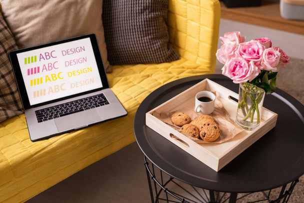 Φορητός υπολογιστής και μαξιλάρια σε κίτρινο καναπέ από μικρό τραπέζι με ξύλινο κουτί που περιέχει δέσμη ροζ τριαντάφυλλα, ποτό και νόστιμα μπισκότα - Φωτογραφία, εικόνα