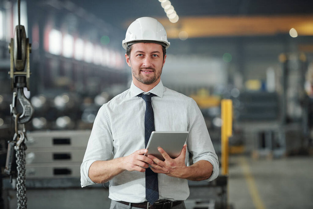 Portrét sebevědomého pohledného technického inženýra v hardhat pomocí tabletu v průmyslové dílně velké továrny - Fotografie, Obrázek