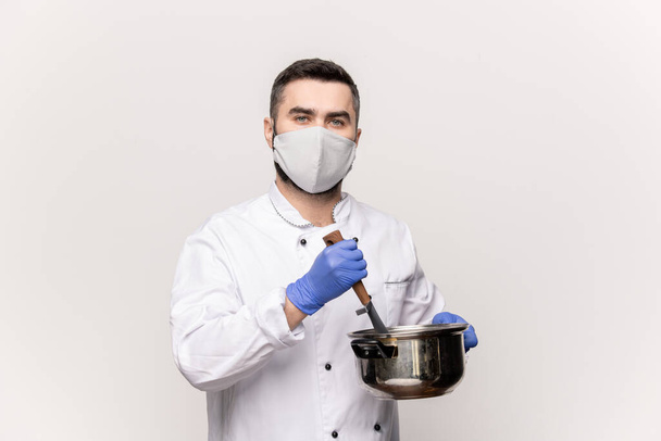 Jovem chef profissional de uniforme, máscara protetora e luvas cortando legumes frescos na panela enquanto cozinha alimentos saudáveis e saborosos
 - Foto, Imagem