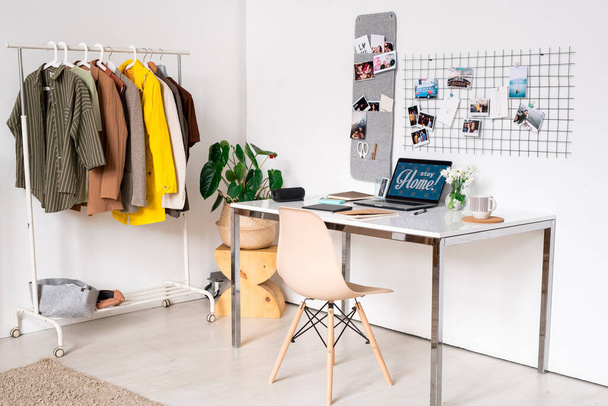Munkahely kortárs divattervező kabátok átadása állvány mentén fal és íróasztal laptop és egyéb kellékek az asztalon - Fotó, kép