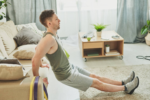 Joven atleta sudoroso en ropa deportiva sosteniendo el sofá a sus espaldas mientras hace ejercicio en la sala de estar durante el período de cuarentena
 - Foto, Imagen