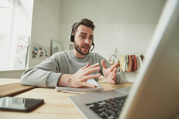 Nuori operaattori kuulokkeilla selittää jotain yhdelle asiakkaalle online-kuulemisen aikana kannettavan tietokoneen edessä kotiympäristössä - Valokuva, kuva