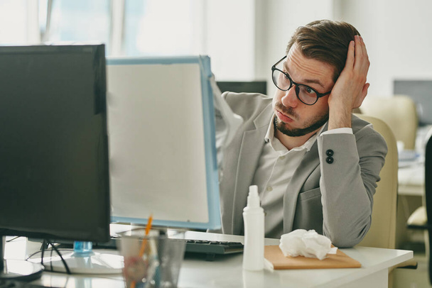 Ärtynyt nuori liikemies silmälaseissa istuu pöydän ääressä ja katselee tietokoneruutua, kun hänellä on työongelmia sepelvaltimoviruksen karanteenin vuoksi - Valokuva, kuva