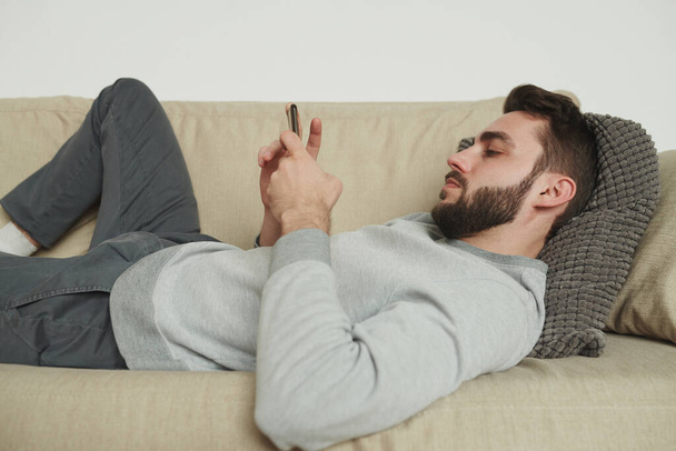 Молодой бородатый мужчина в трениках и толстовке расслабляется на диване в гостиной и прокручивается в смартфоне во время самоизоляции
 - Фото, изображение
