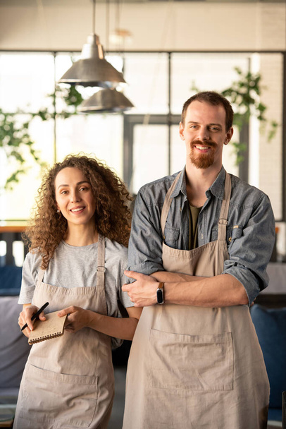 Two young cheerful owners of contemporary restaurant or cafe standing in front of camera and waiting for new guests in the morning - Photo, Image