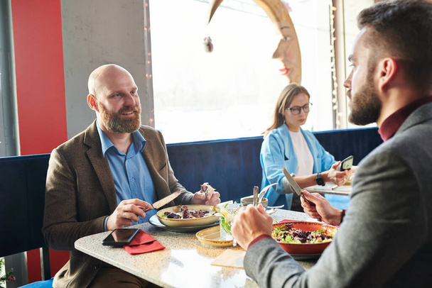 Uśmiechnięty łysy biznesmen siedzący przy stole i jedzący sałatkę podczas rozmowy z kolegą podczas lunchu w kawiarni - Zdjęcie, obraz