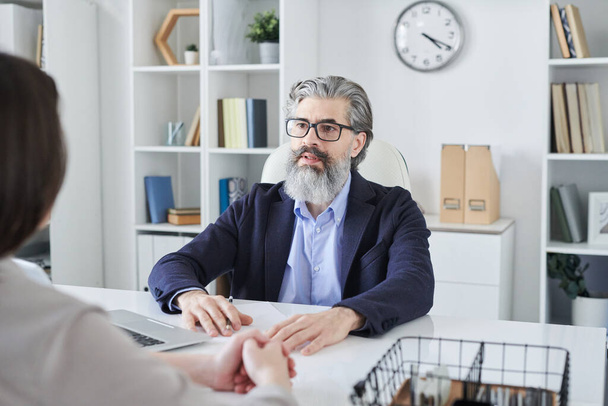 Starší vousatý muž profesionál v brýlích a formalwear při pohledu na svého klienta během konzultace nebo setkání v kanceláři - Fotografie, Obrázek