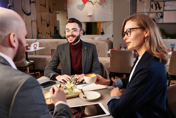 飲食店でコーヒーを飲みながら、ガジェットとテーブルに座ってプロジェクト実施の戦略を議論する若いビジネスの人々 - 写真・画像