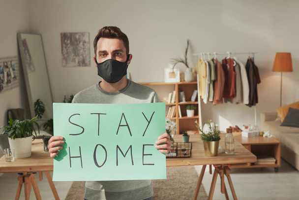 Młody chory mężczyzna w czarnej masce na twarzy trzymający plakat z rekomendacją zostania w domu i pokazywania go tobie. - Zdjęcie, obraz