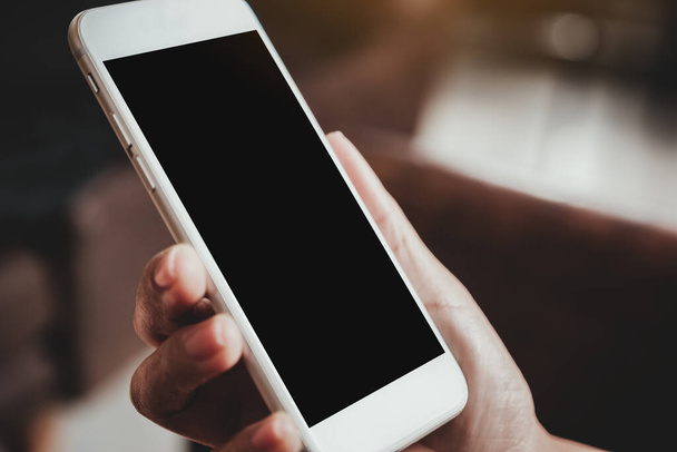 Εικόνα mockup ενός χεριού που κρατά λευκό κινητό τηλέφωνο με κενή μαύρη οθόνη επιφάνειας εργασίας με θολό φόντο - Φωτογραφία, εικόνα