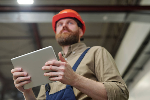 Debajo de la vista del joven ingeniero barbudo en hardhat usando tableta mientras controla el trabajo en fábrica
 - Foto, imagen