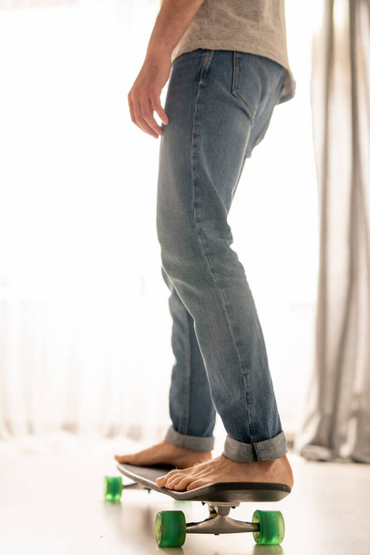 Nierozpoznawalny mężczyzna w dżinsach uczy się jeździć na desce po salonie w izolacji domowej - Zdjęcie, obraz