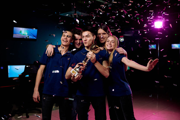Grupa pięciu nastolatków świętuje zwycięstwo w zawodach cybersportu stojąc w upadku konfetti i pokazując złoty puchar - Zdjęcie, obraz