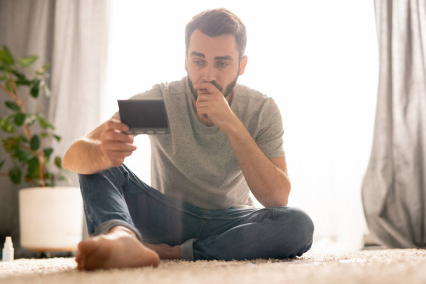 Poważny zamyślony młody człowiek siedzi na dywanie i pochyla głowę w dłoni patrząc na fotografię i zaginione stare życie przed koronawirusem - Zdjęcie, obraz