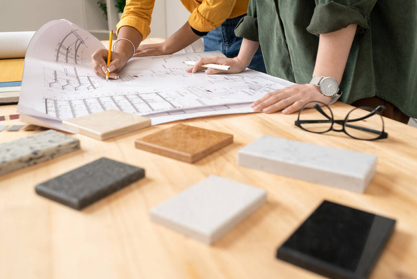 Руки двух молодых дизайнеров интерьера, стоящих за столом и наклоняющихся над планом квартиры, пока один из них рисует
 - Фото, изображение