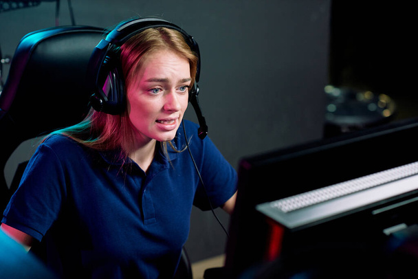 Young female e-sport addict in headset looking at computer screen during cyber competition while sitting in modern game club - Photo, Image