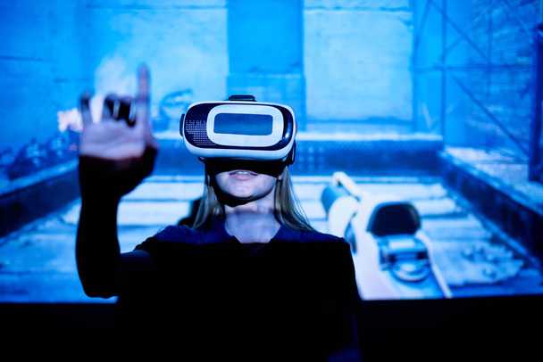 Mujer joven con gafa vr pulsando el botón virtual mientras está de pie contra la pantalla grande con la proyección de un nuevo videojuego e-sports
 - Foto, Imagen