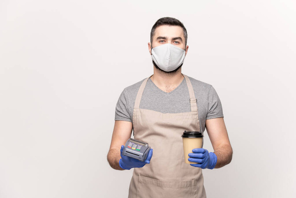 Young waiter in workwear, protective mask and gloves holding your drink and payment machine while standing against white background - Photo, Image