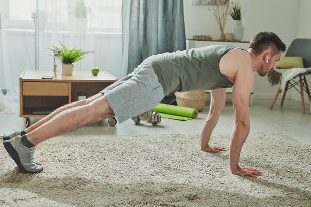 Young man in sportswear and earphones doing press-ups on the floor of living-room at home during period of quarantine - Photo, Image
