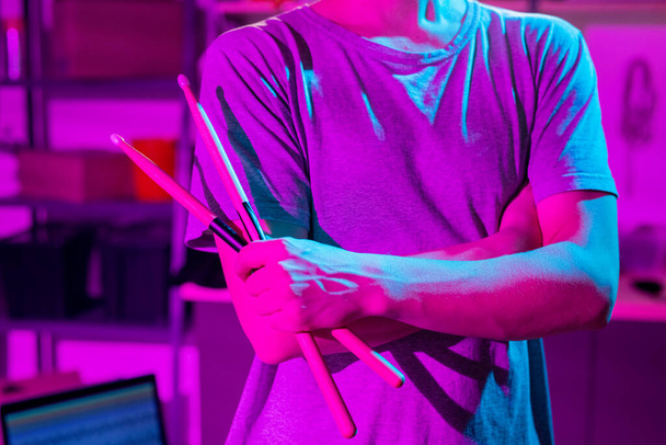 Parte média do jovem em t-shirt segurando baquetas enquanto cruza os braços pelo peito durante o ensaio com tambores e címbalos na garagem
 - Foto, Imagem