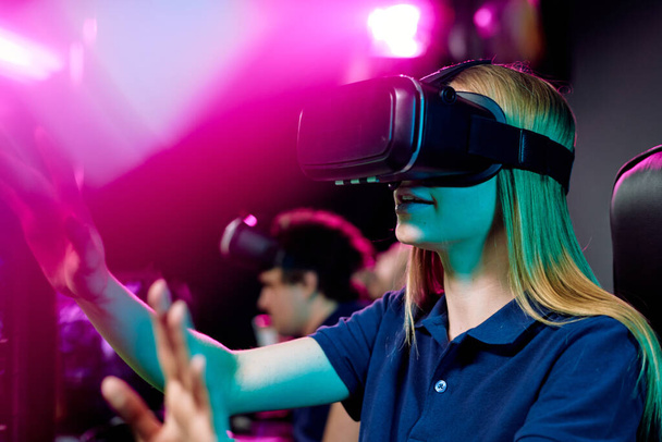 Blond teenage girl in vr goggle touching virtual display while taking part in cybersports video game on background of her friends - Фото, изображение