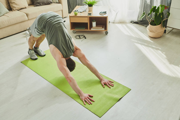 Junger Sportler in Aktivkleidung beugt sich beim Training im Wohnzimmer vor, während er mit Füßen und Händen auf Matte auf dem Boden steht - Foto, Bild